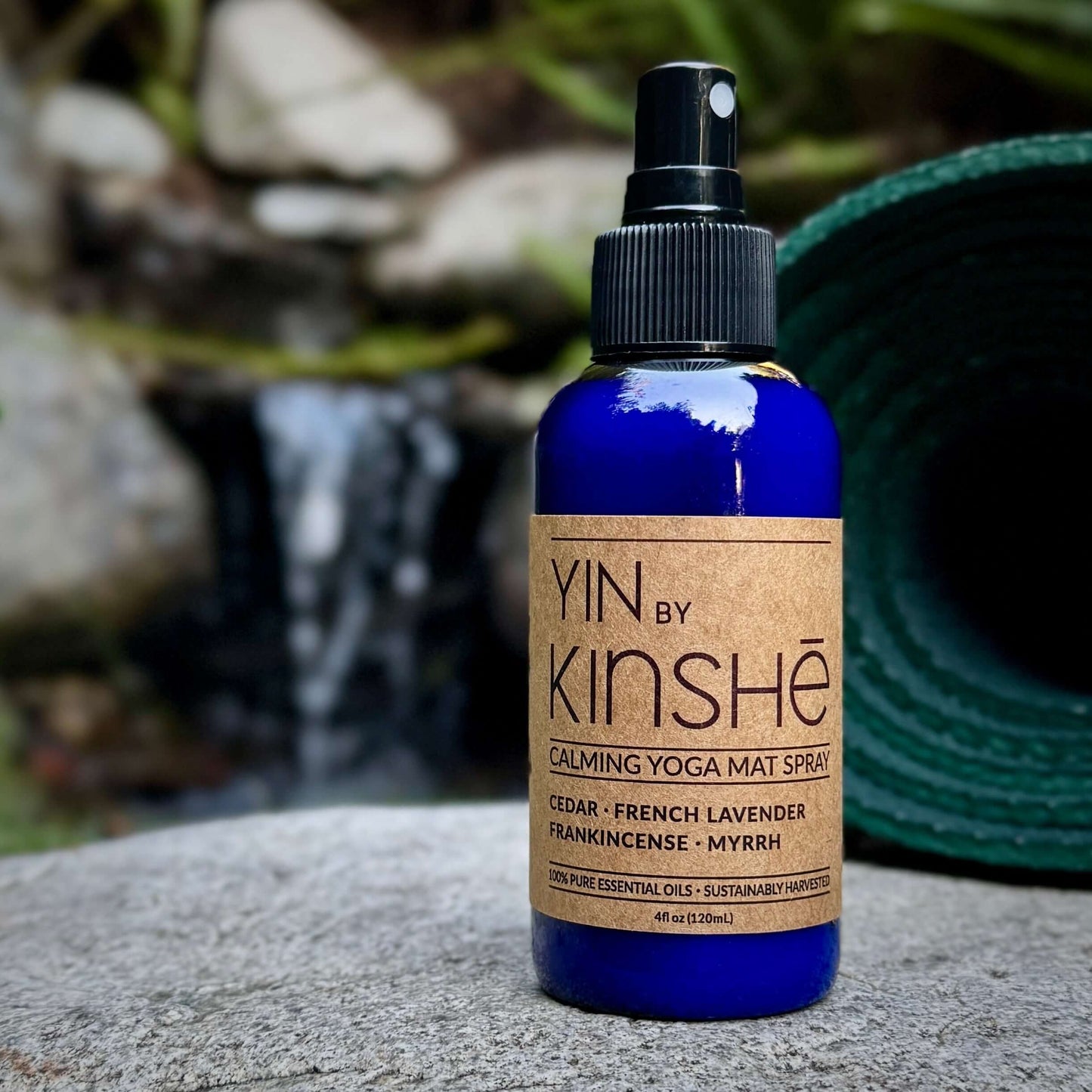 Sapphire blue bottle of calming mat refresh spray stands on smooth stone near a little waterfall. The label reads "Yin".