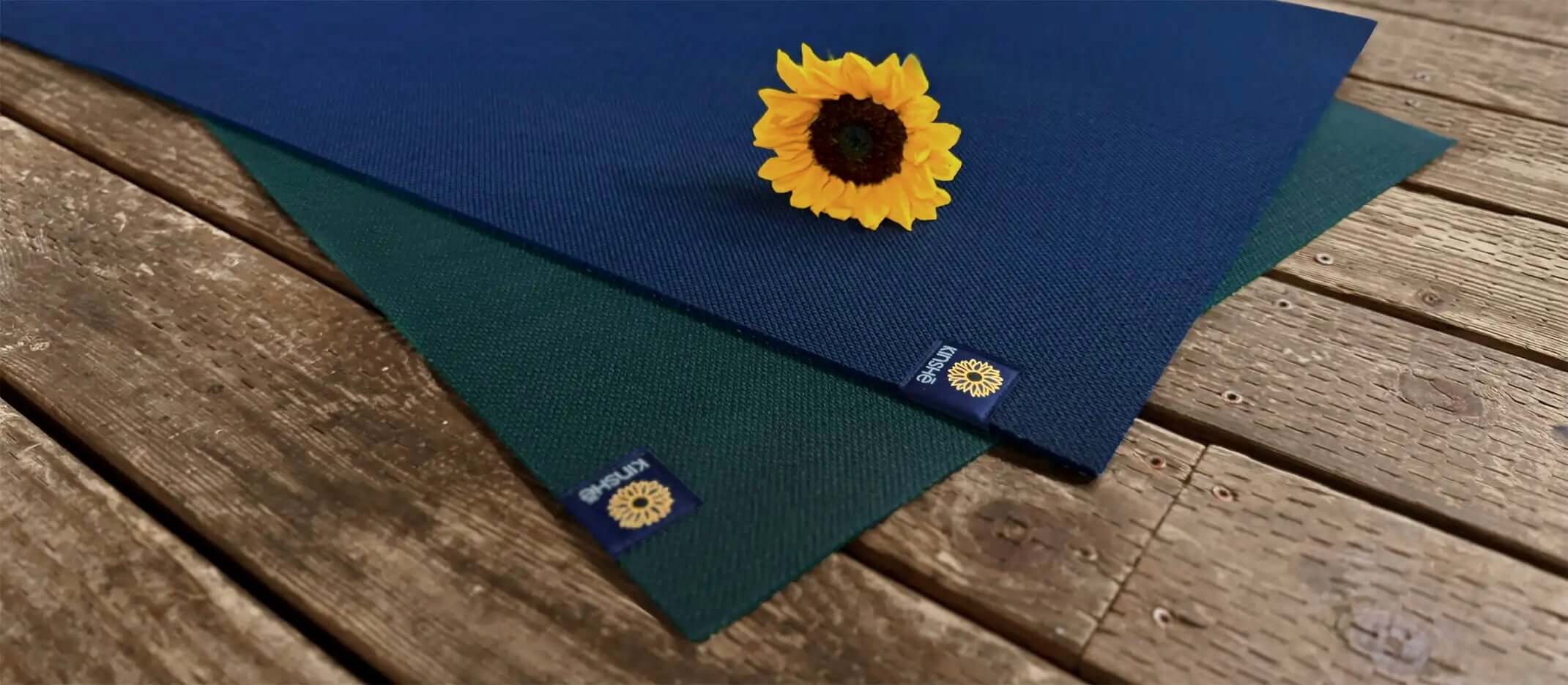 A sunflower lies on two Kinshē yoga mats fanned out on wood deck. Mat colors are Neptune blue and jewel forest green.