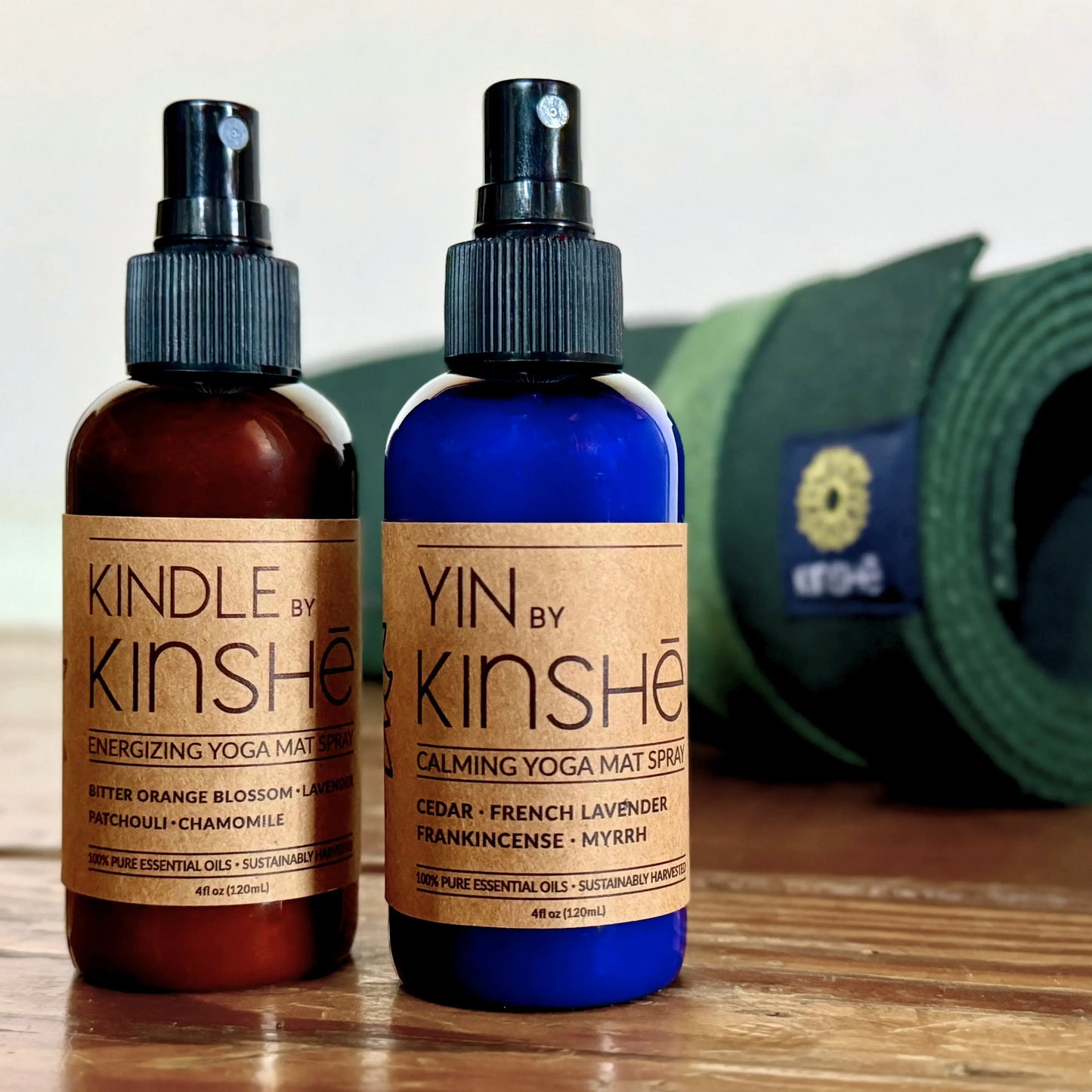 A blue bottle of calming mat spray sits next to an amber bottle of energizing mat spray in front of a green yoga mat.