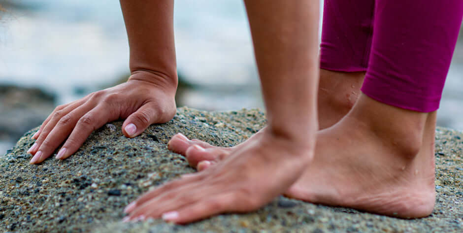 Close up on a woman’s hands and feet as she stands in forward bend on a rocky shoreline.