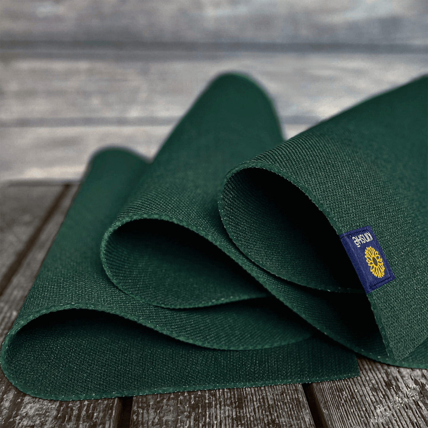 A forest green Kinshē Yoga Earthmat, a non-slip TPE yoga mat, unspools on a wood deck — sustainably crafted and guaranteed for life.
