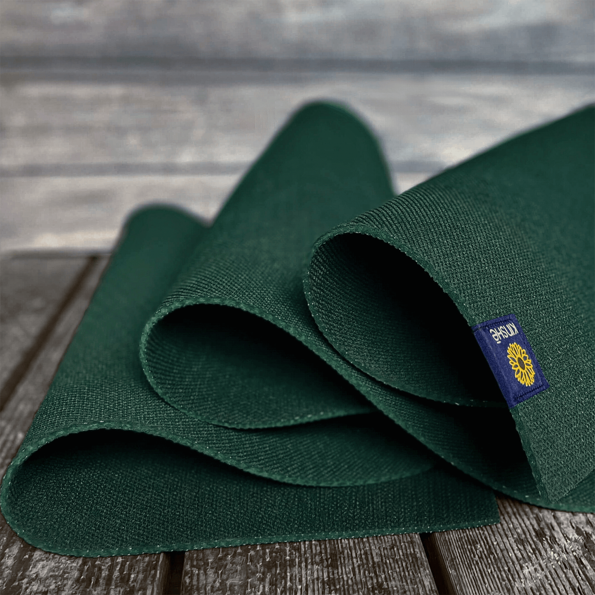 A forest green Kinshē Yoga Earthmat, a non-slip TPE yoga mat, unspools on a wood deck — sustainably crafted and guaranteed for life.