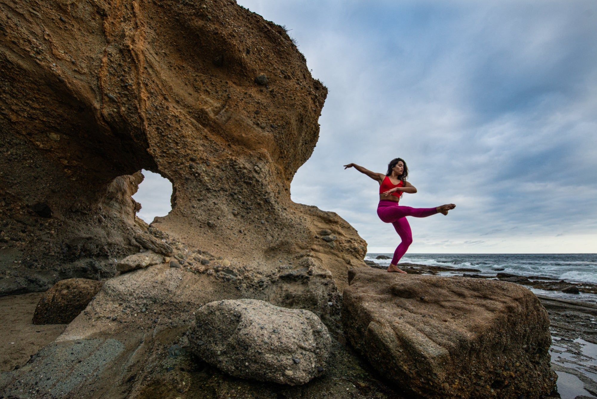 A yogini next to a sea cave balances on one leg with the other leg extended.