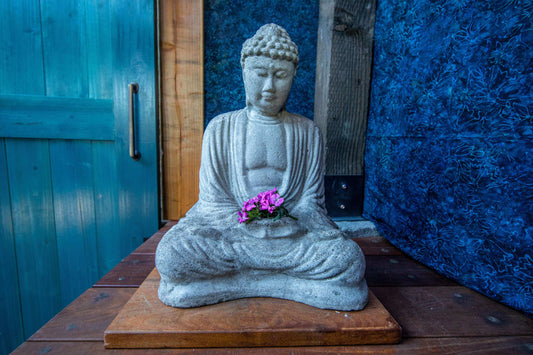 Purple flowers are arranged in the lap of a  grey stone buddha with a blue background.  