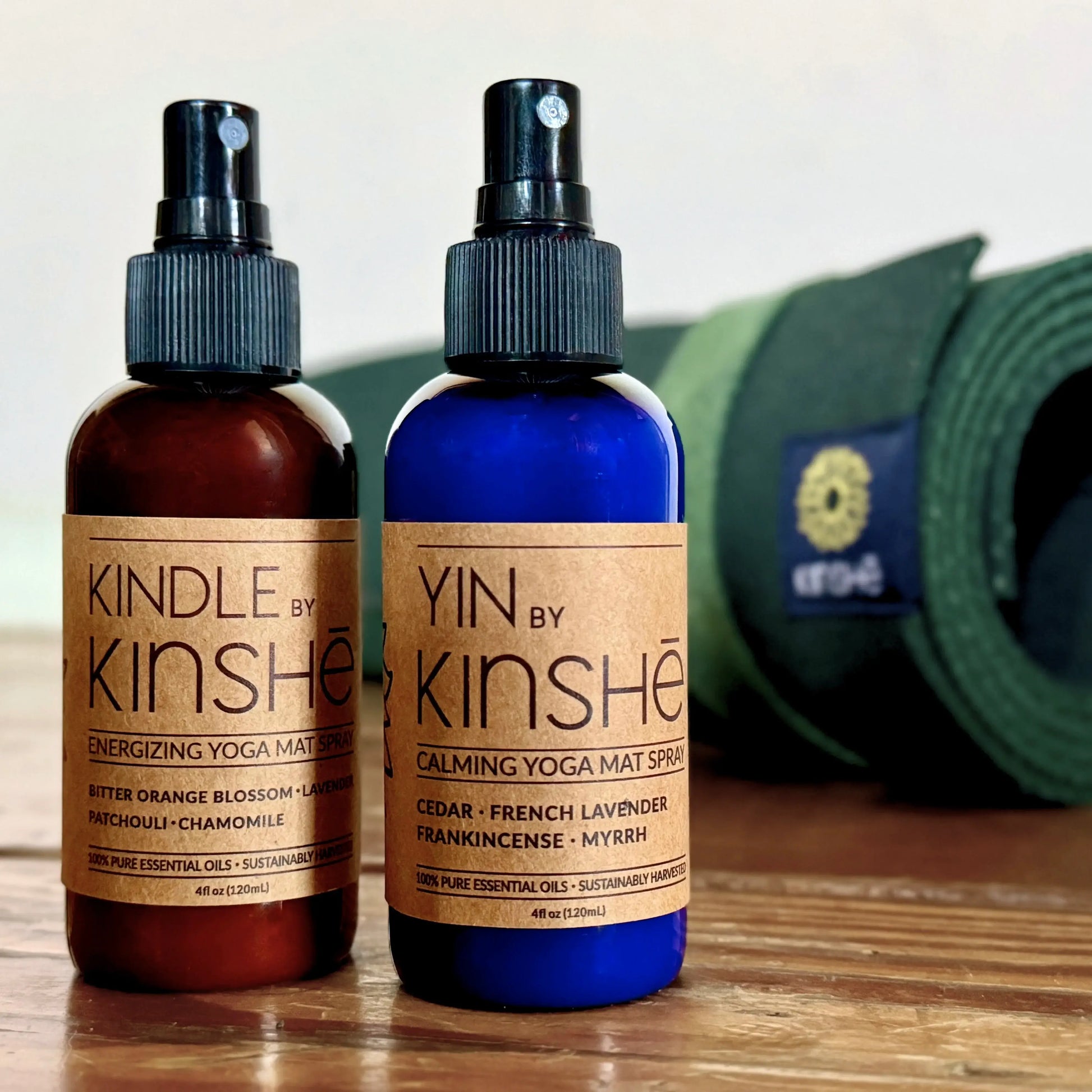 A blue bottle of calming mat spray sits next to an amber bottle of energizing mat spray in front of a green yoga mat.