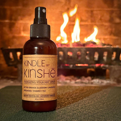 An amber bottle of Kindle energizing yoga mat refresh spray stands near a cozy fire.