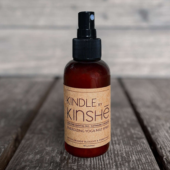 A bottle of KINDLE natural yoga mat spray with energizing pure essential oils stands on a grey wood deck