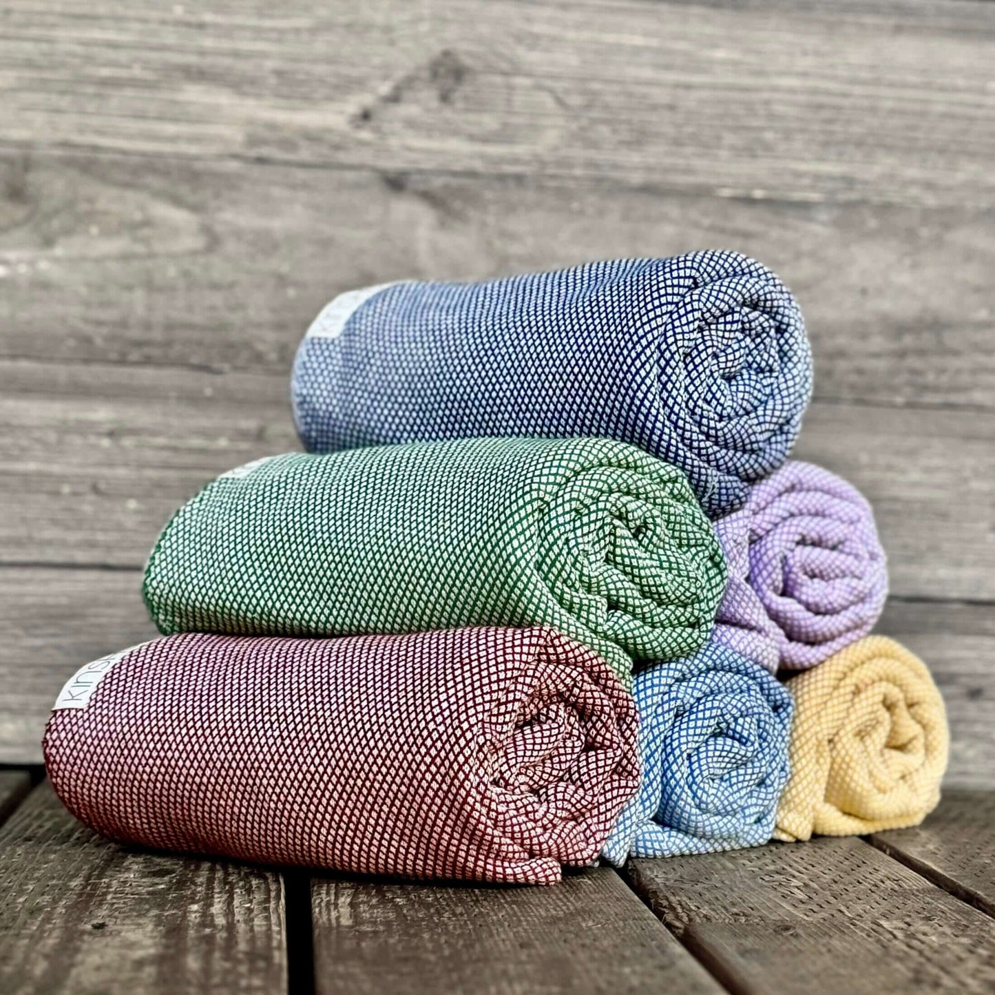 Rolled Turkish cotton yoga towels in Neptune blue, forest green, turmeric and garnet stacked in a pyramid on wood deck.